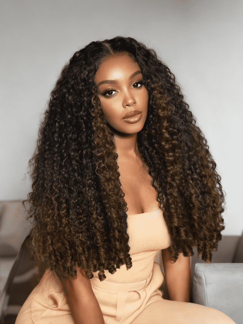 A model displays the UNice Stylish Golden Highlights Add Depth To Body Wave Texture&&Human Hair Wig & Can, showing a woman sitting on a couch with long curly hair.