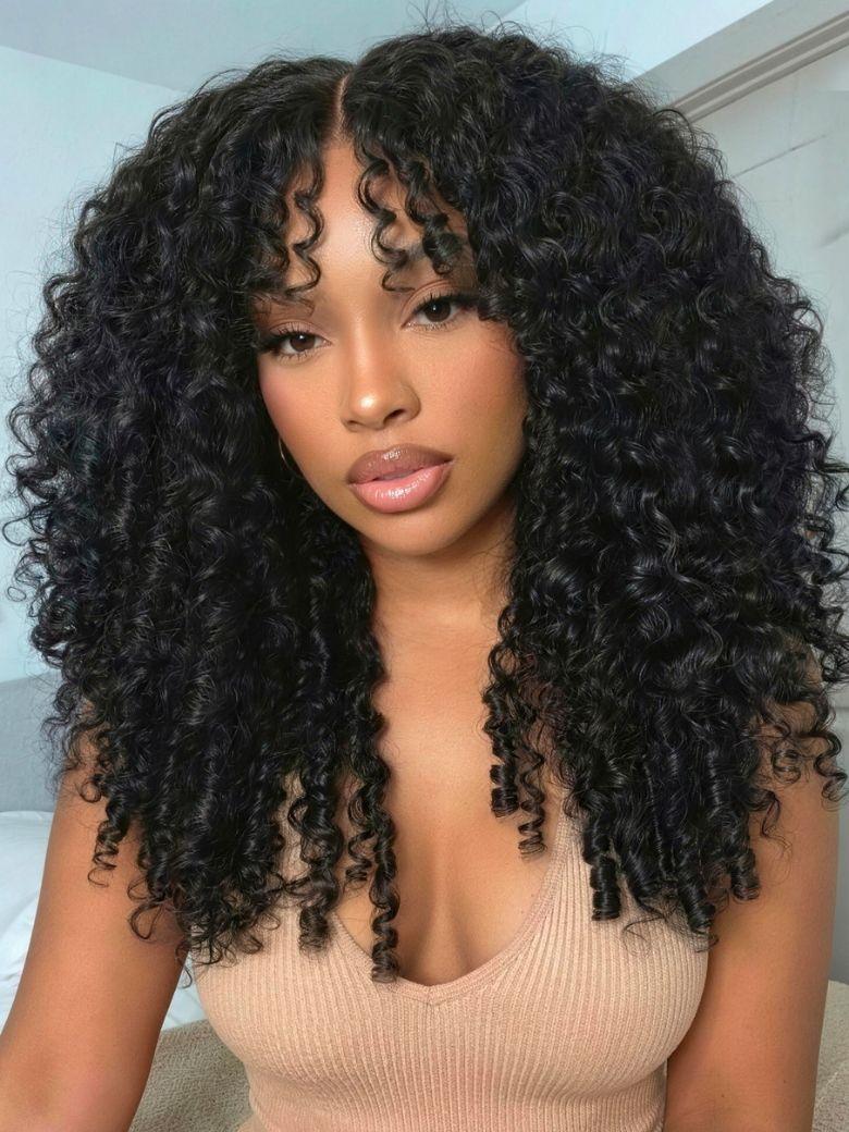 A woman with voluminous, defined curly hair and restyled wispy bangs poses indoors, showcasing a gorgeous Burmese curly wig. She wears a beige top and natural makeup.