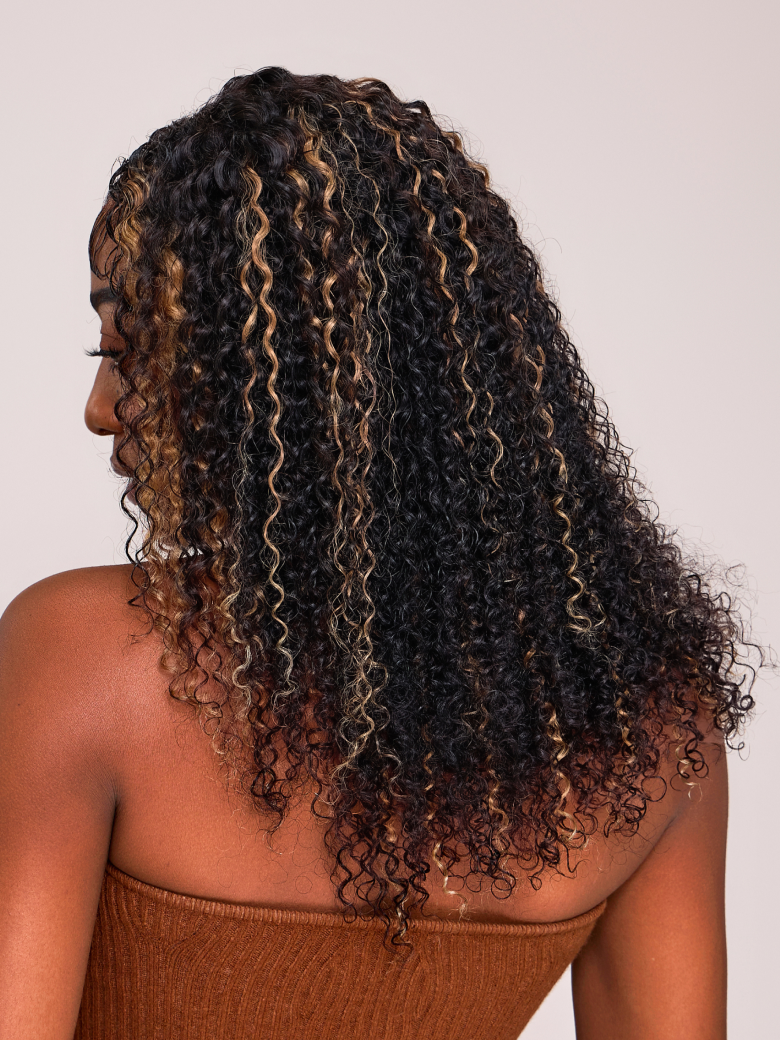 A model displays the UNice 6x4.5 Pre Cut Lace Curly Wig Put On And Go Quick Easy Black With Blonde Highlights, showing a model with long curly hair wearing a brown top.